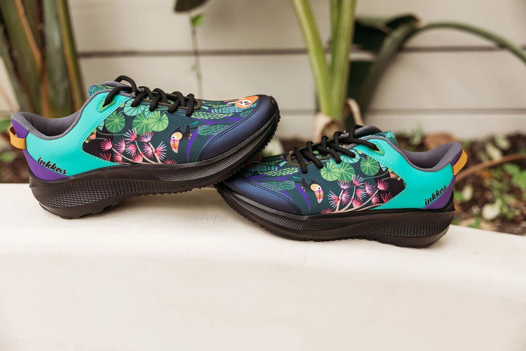 A pair of low-top athletic sneakers with a vibrant tropical floral print, resting on a white ledge with a blurred garden background.