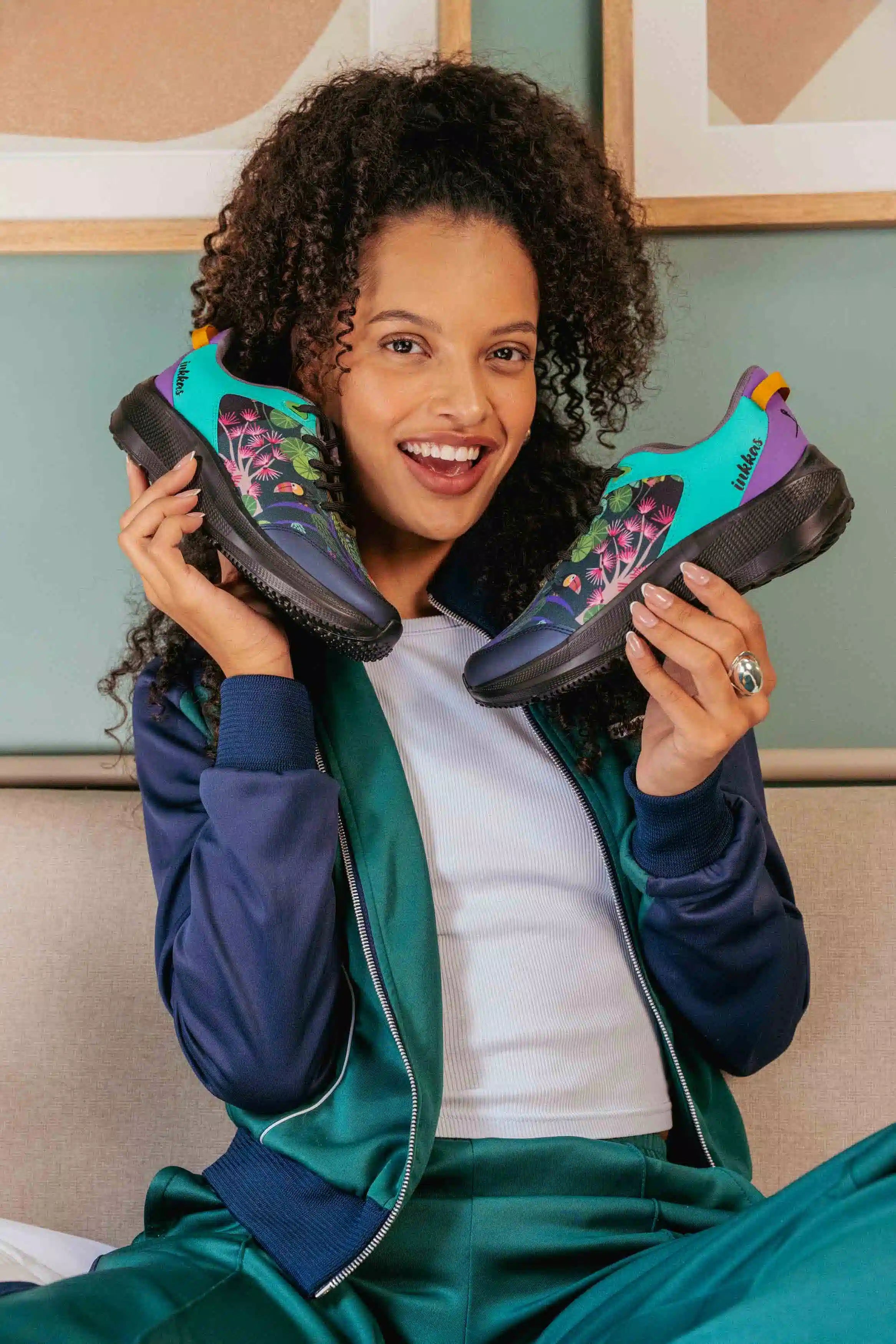 A smiling woman with curly hair holds up a pair of colorful athletic sneakers, wearing a navy and teal satin tracksuit over a white t-shirt in an indoor setting.