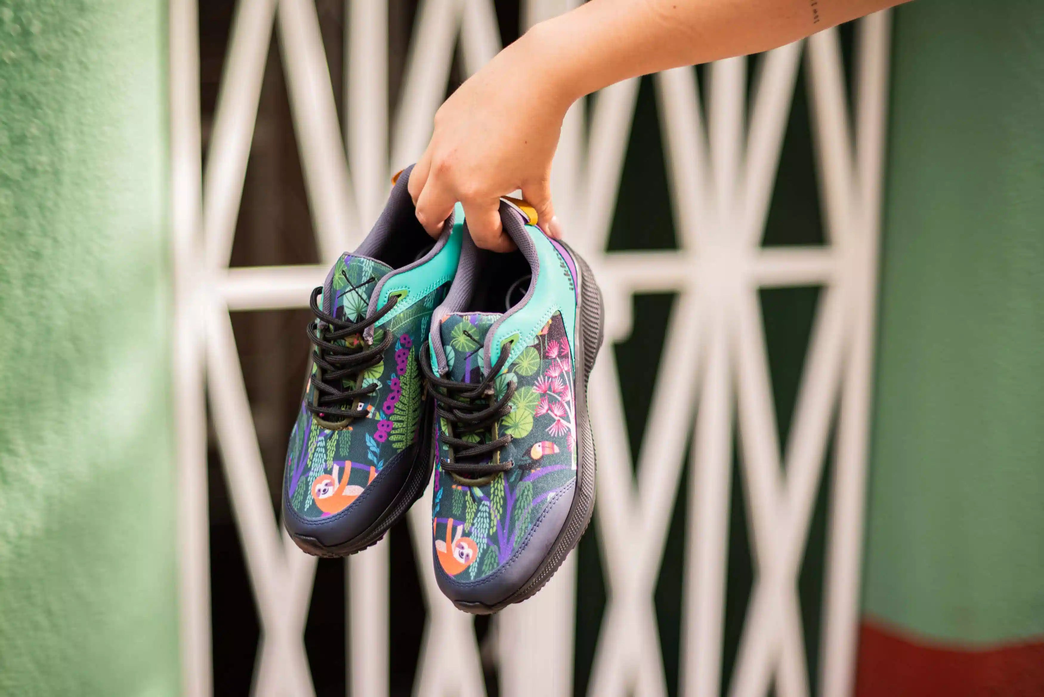 A pair of colorful children's sneakers with a jungle-themed print, held by a hand in front of a white metal gate and green wall.