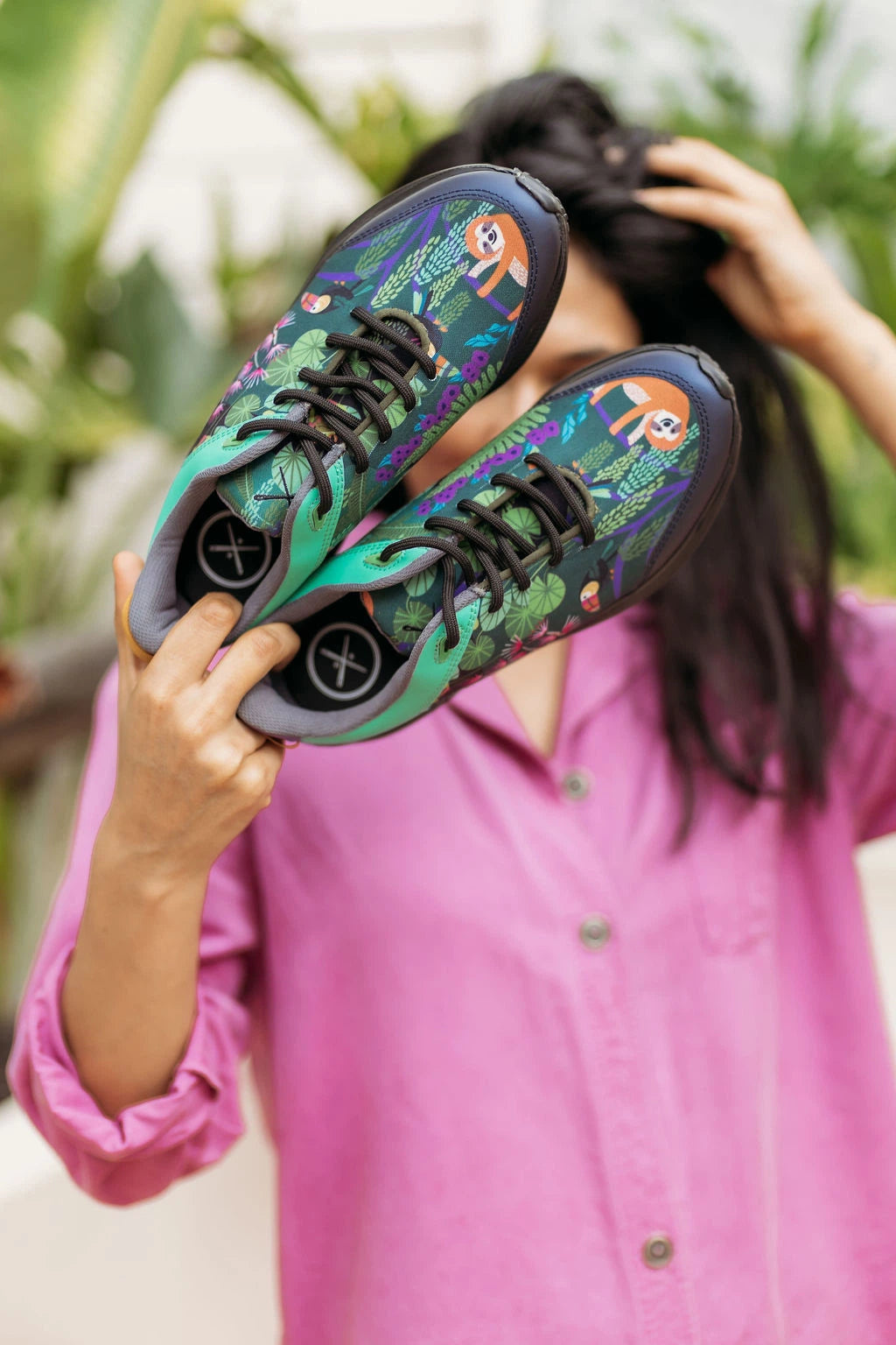 A person in a pink shirt holds up a pair of colorful sneakers with a tropical floral pattern featuring sloths and green leaves. The background is a soft-focus outdoor setting with green foliage.