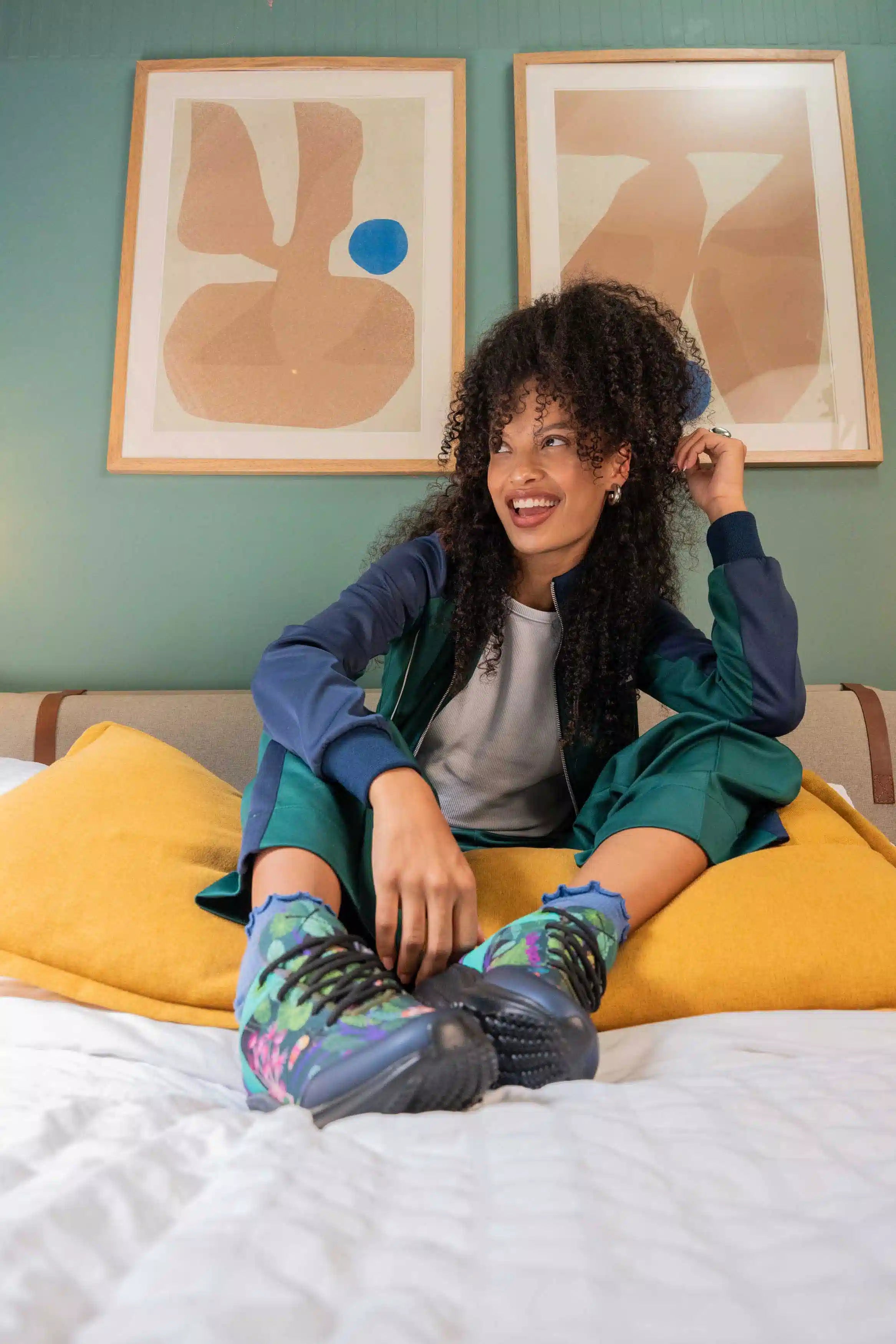 A woman sits on a bed wearing a matching teal and navy tracksuit, a gray t-shirt, and colorful patterned sneakers. She is posed in front of two framed abstract art prints on a sage green wall.