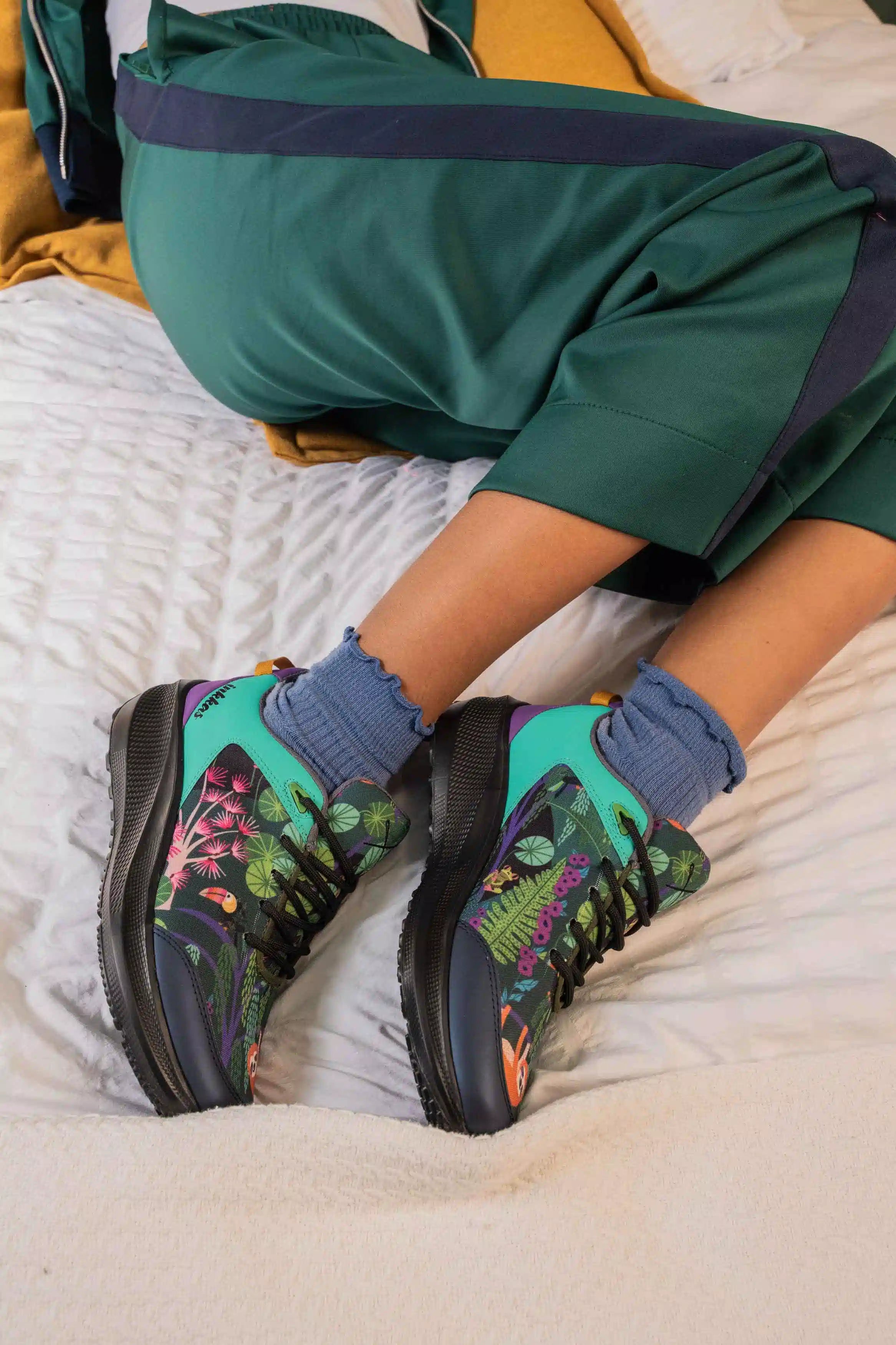 A person reclining on a bed wearing green tracksuit pants and colorful athletic sneakers with blue socks. The sneakers feature a vibrant tropical leaf print in green, pink, and purple on a teal and black base.