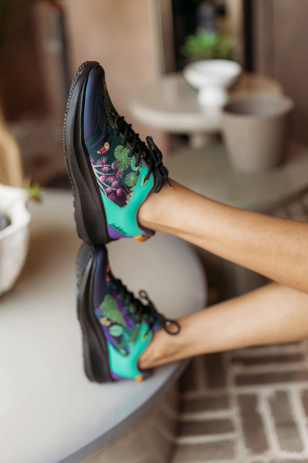 A person's legs crossed in a modern indoor setting, wearing a pair of colorful athletic sneakers with a black base and a vibrant turquoise, green, and purple floral pattern on the sides.
