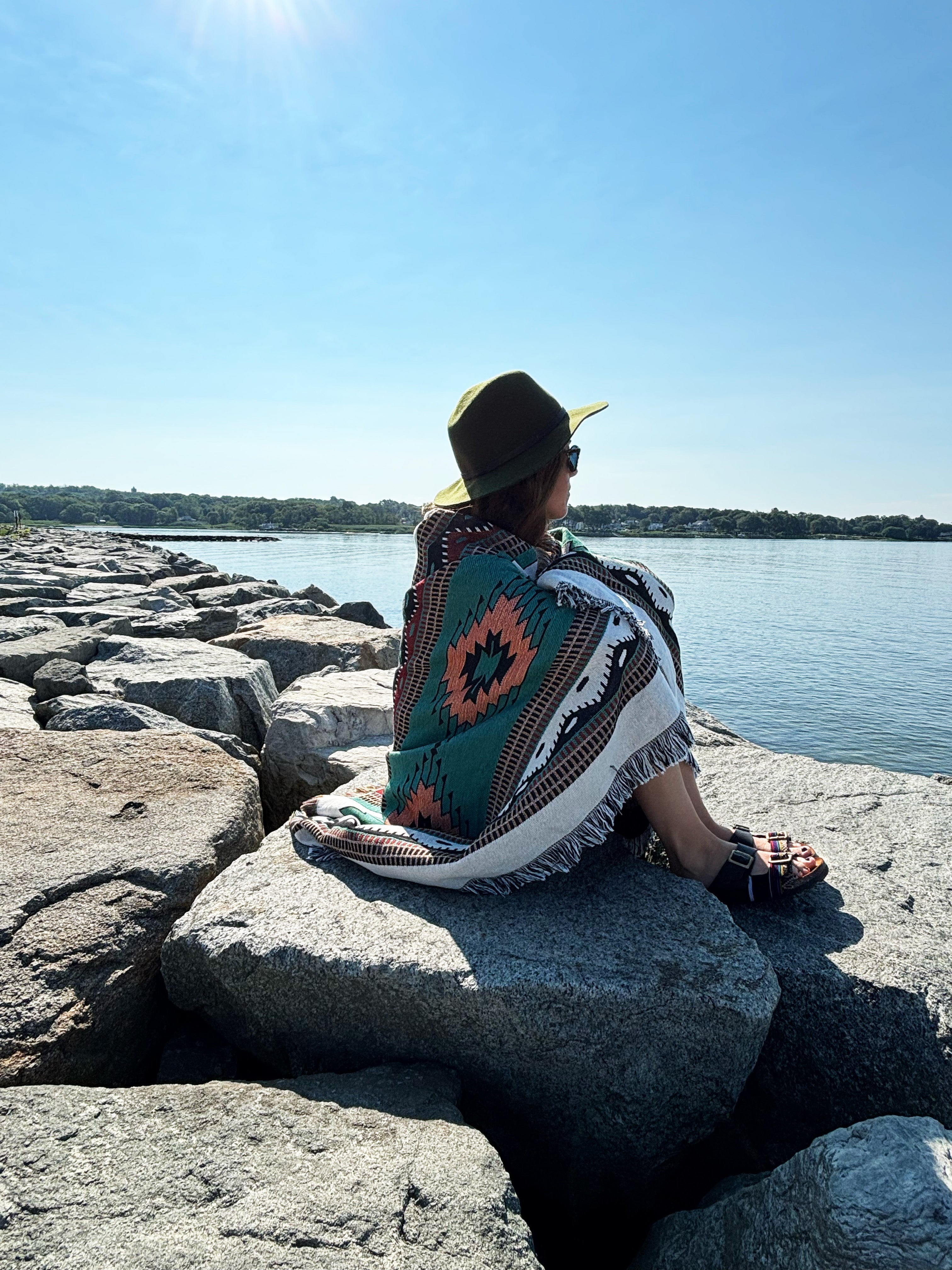 Person sitting on rocks by a body of water, wearing a colorful patterned blanket and hat.