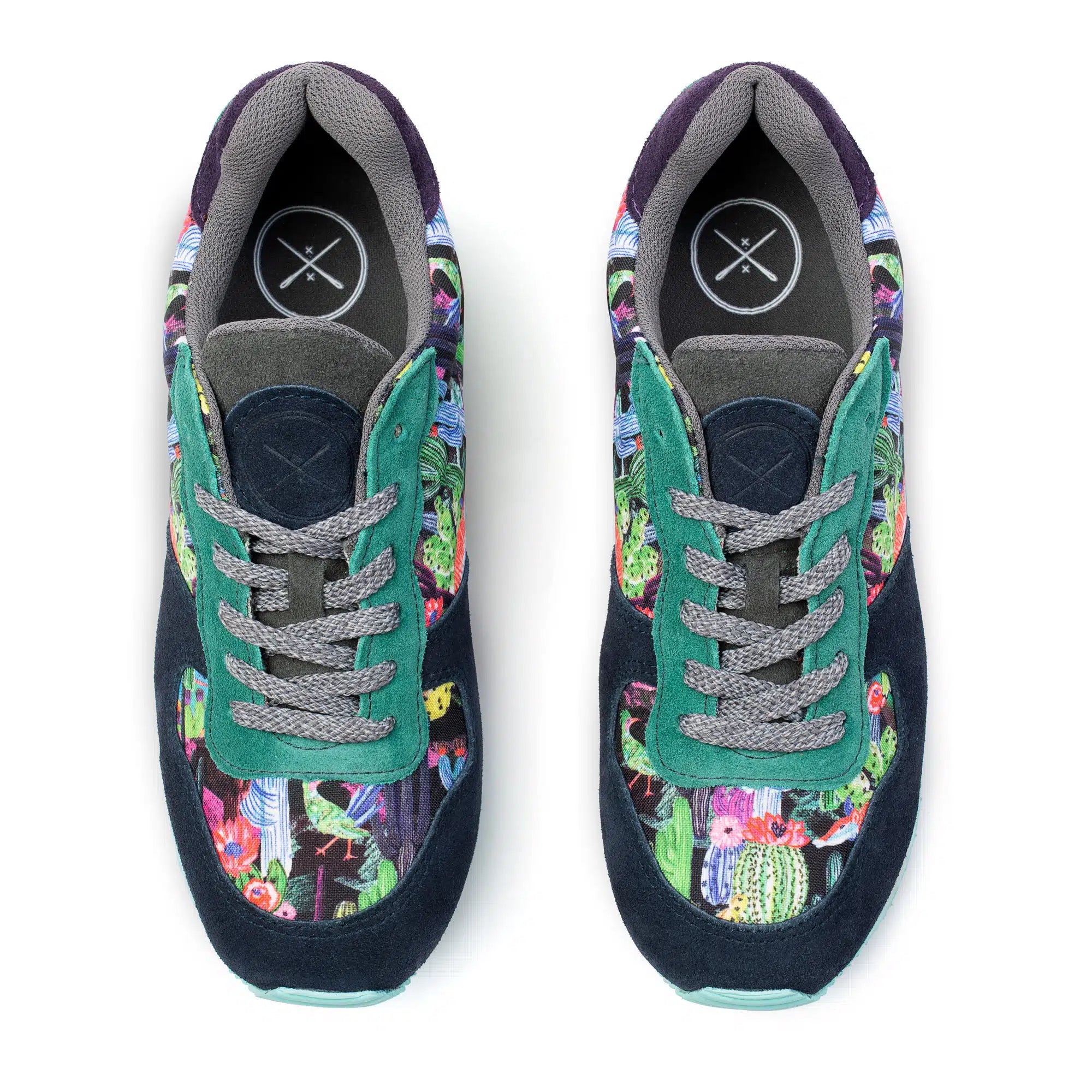 A pair of low-top sneakers shown from a top-down view on a white background. The shoes feature a dark navy suede base with green suede overlays, gray laces, and a colorful, busy floral and botanical print on the sides.