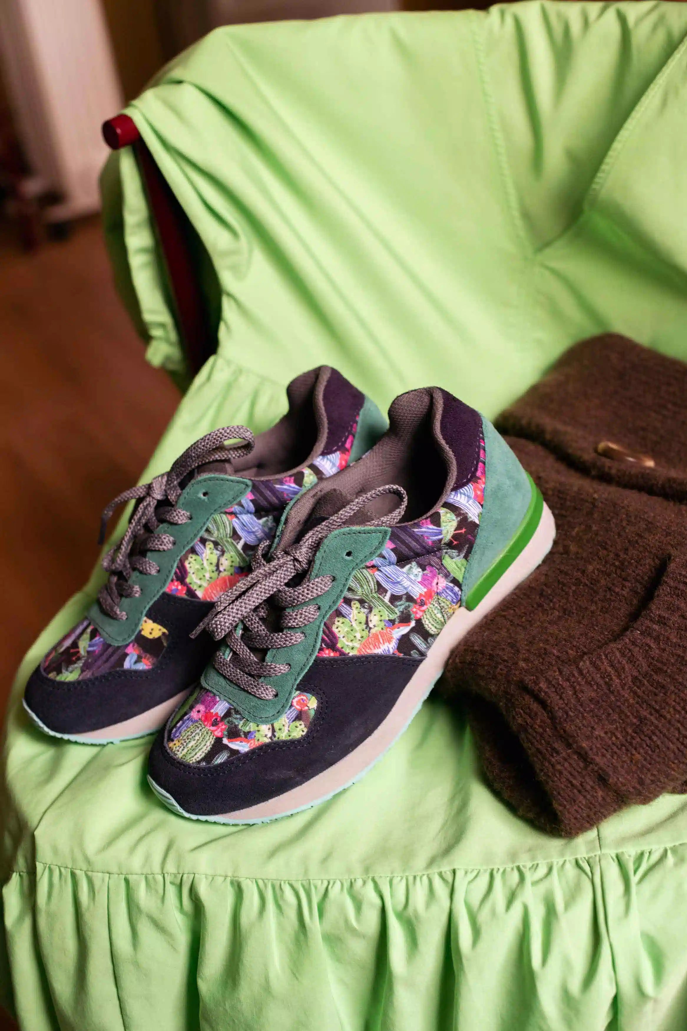 A pair of low-top sneakers with a dark navy base, green suede overlays, and a colorful floral print on the sides, styled on a lime green chair next to a brown knit sweater.