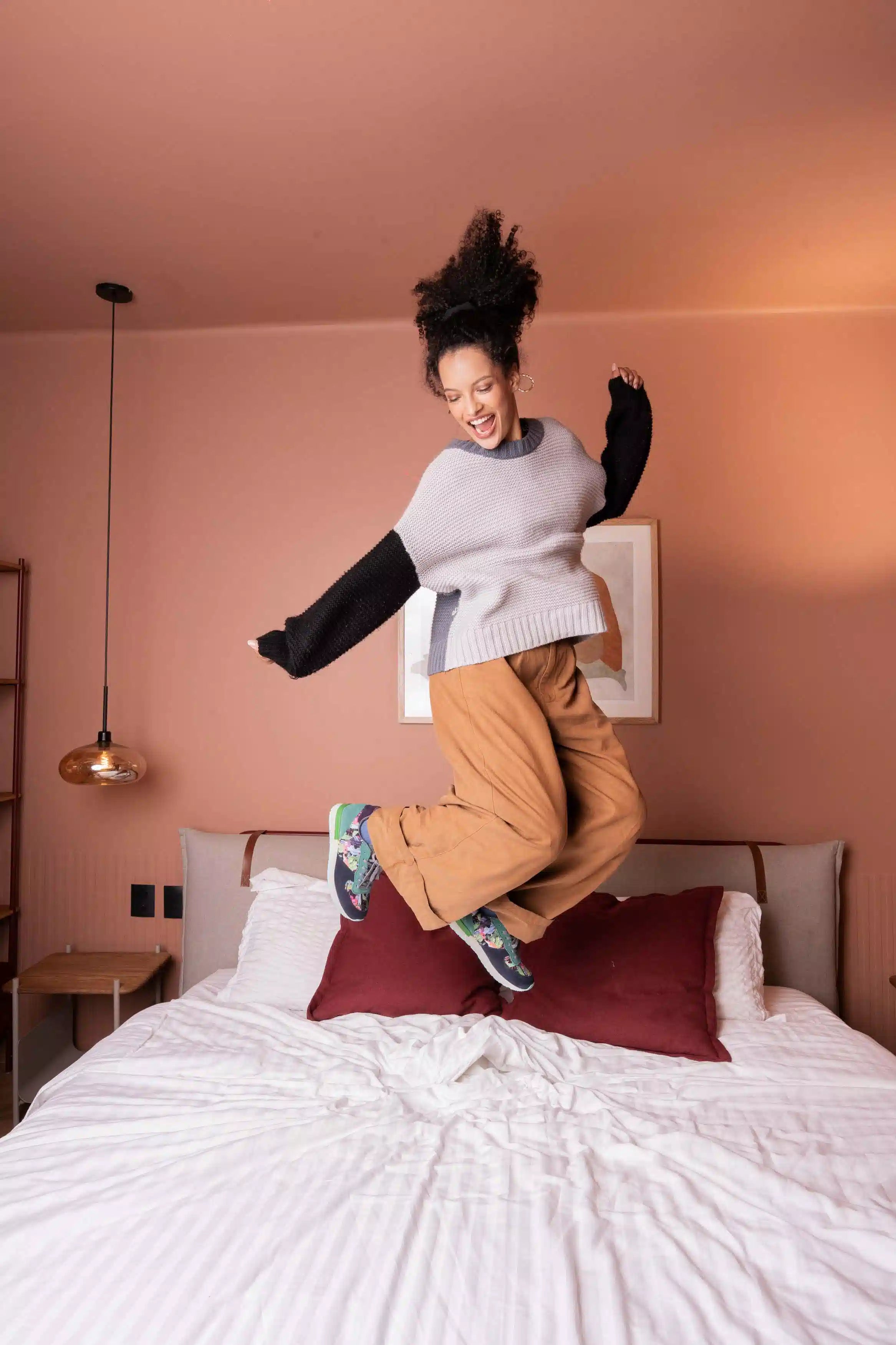 A woman jumps excitedly on a bed in a bedroom with dusty pink walls. She is wearing a light gray knit sweater over a black long-sleeve shirt, loose tan pants, and colorful sneakers.