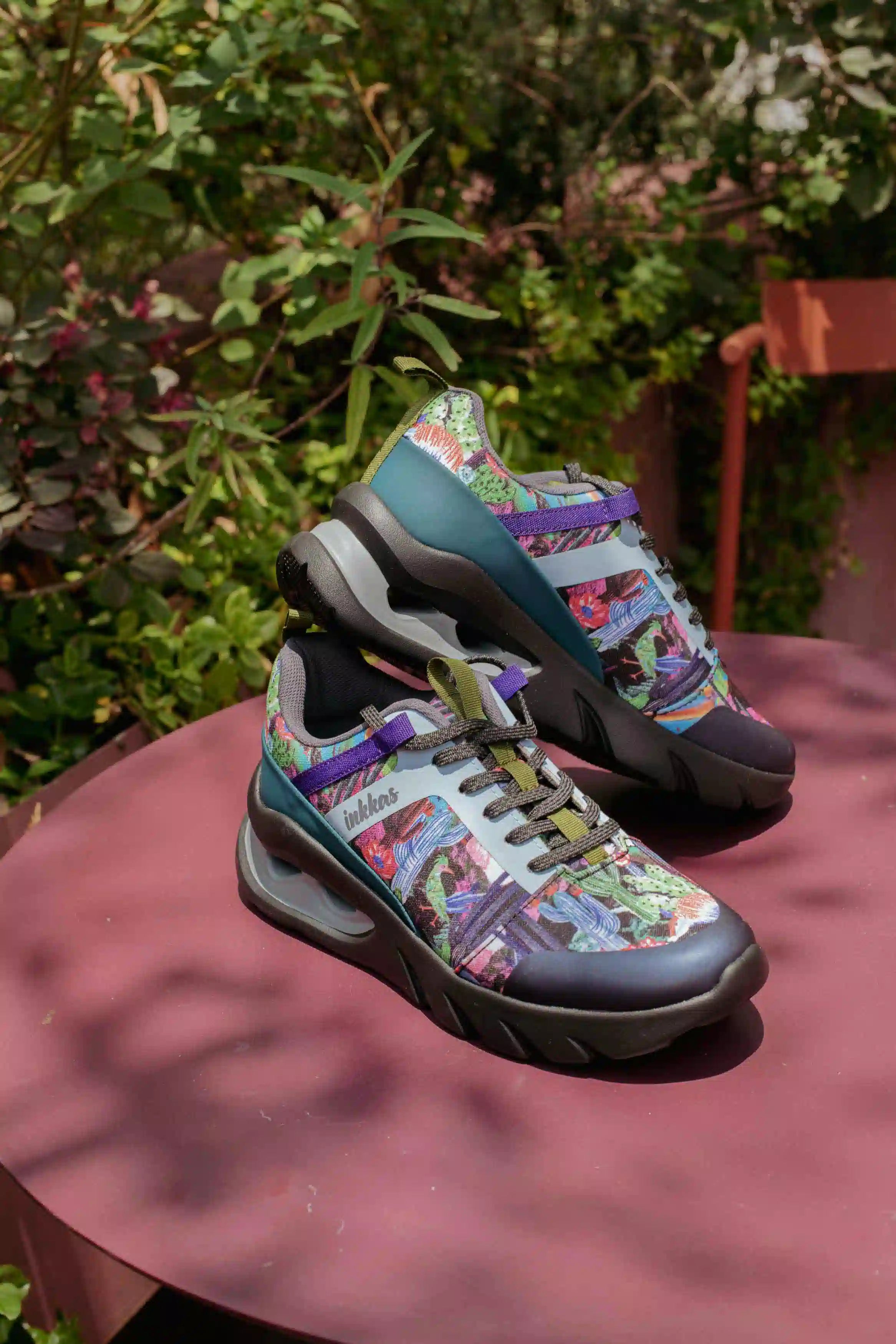 A pair of colorful, highly patterned athletic sneakers displayed on a dusty rose table outdoors with green foliage in the background.