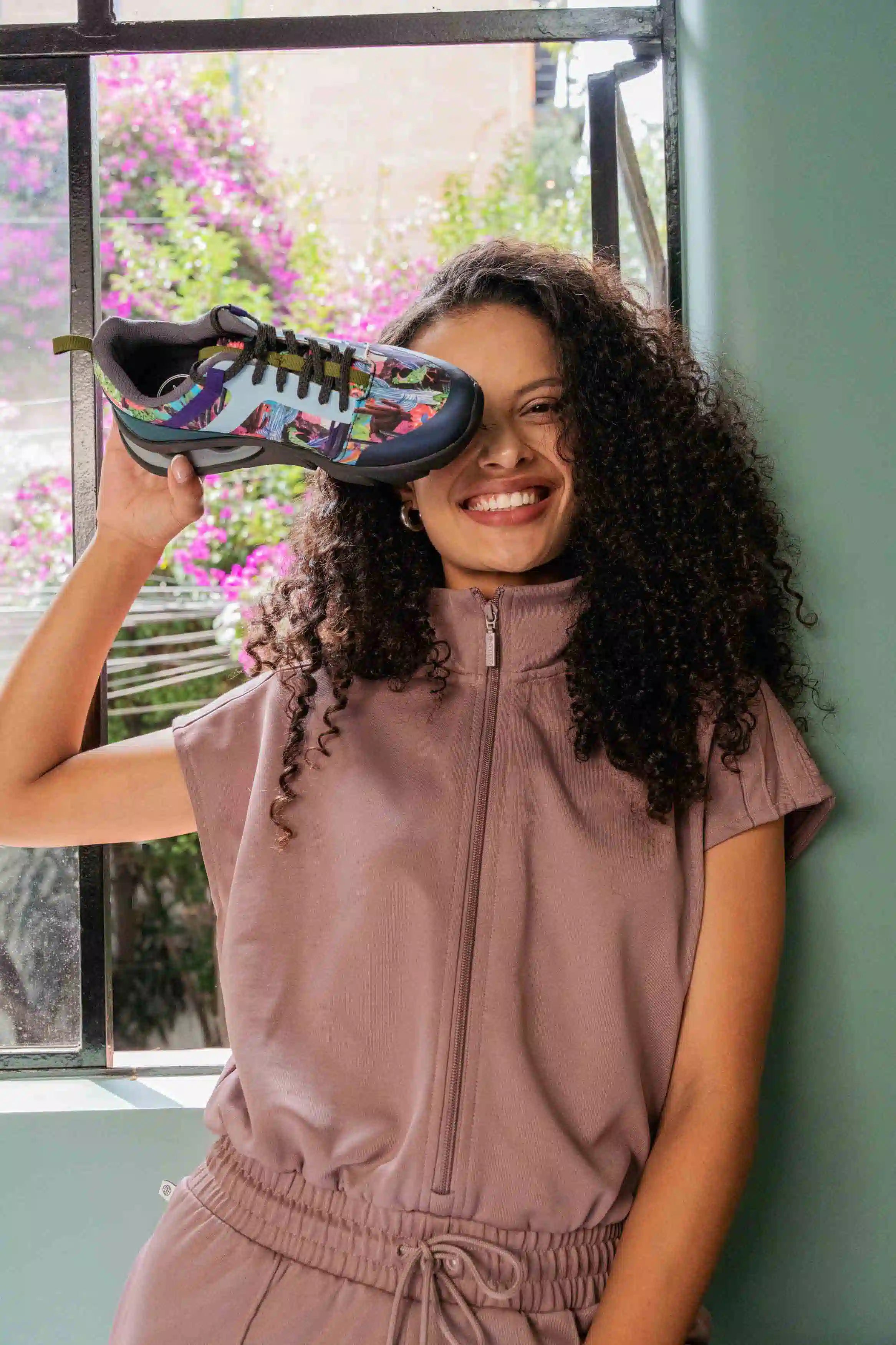 A smiling woman with curly hair holds up a colorful athletic sneaker to show the sole, wearing a mauve zip-up tracksuit in front of a window with pink flowers outside.