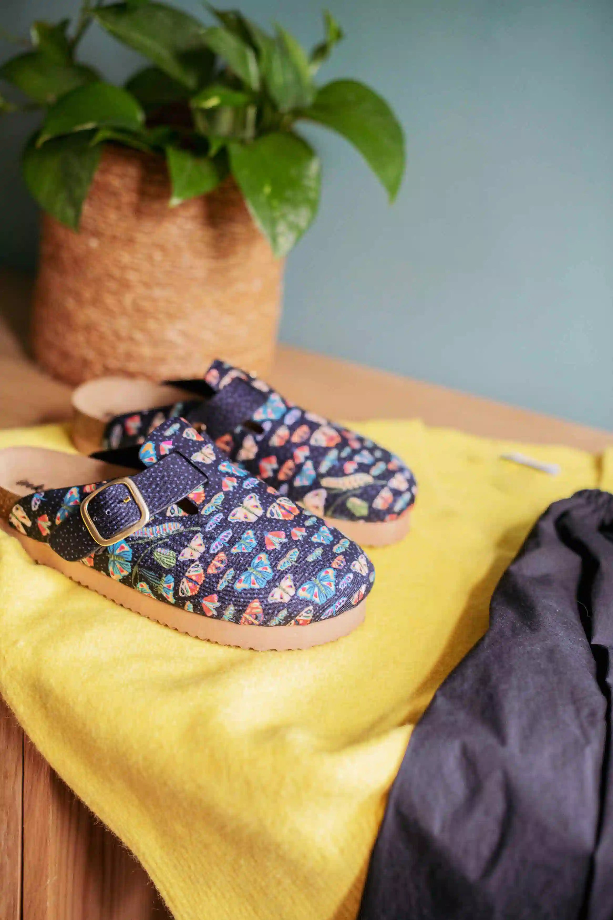 A pair of slip-on clogs with a colorful fish pattern on a navy blue background, styled on a yellow blanket next to a dark gray fabric, with a potted green plant in the background.