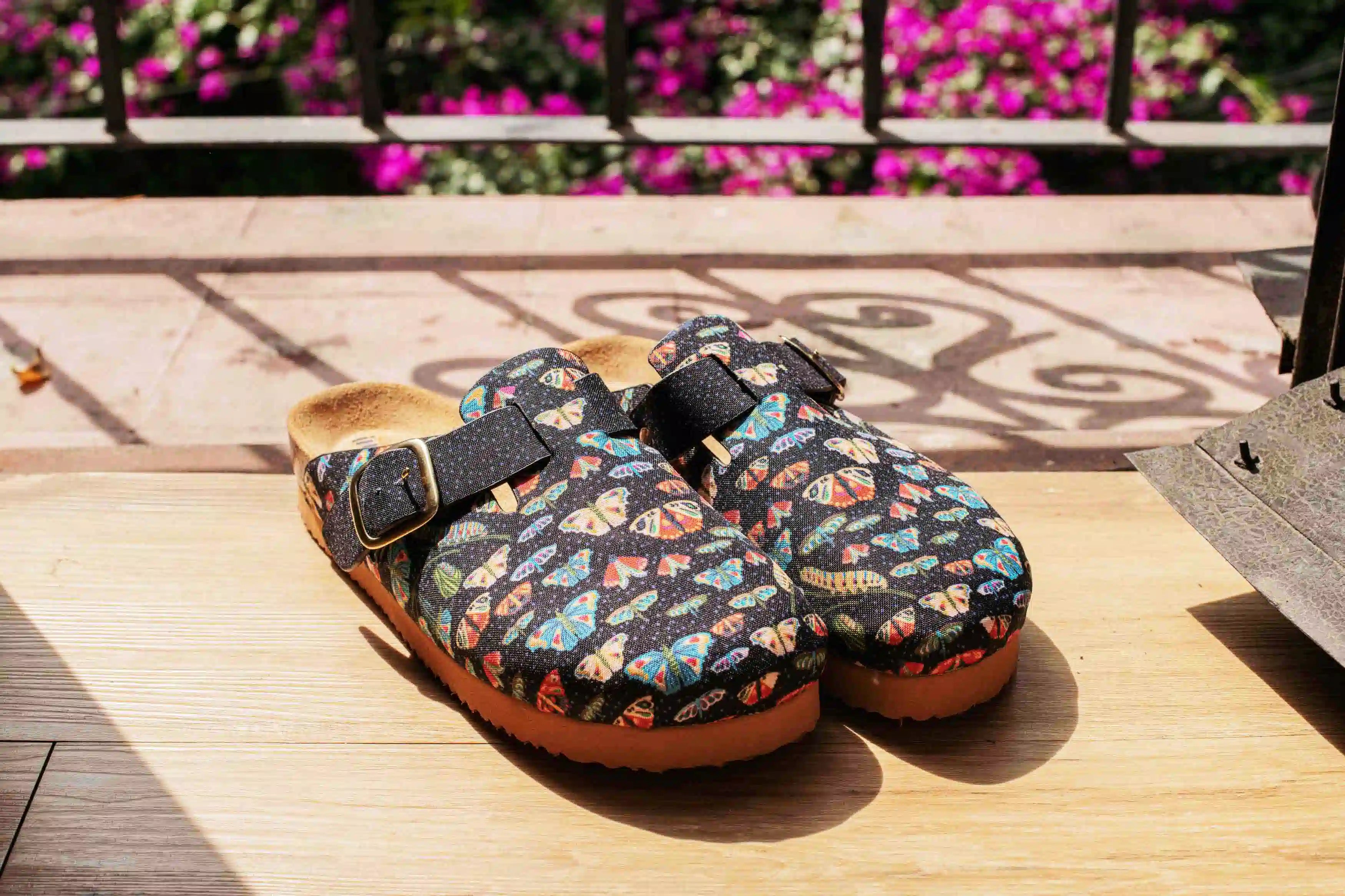 A pair of clogs with a colorful butterfly pattern on a black background, resting on a wooden deck with a blurred background of flowers and a metal railing.