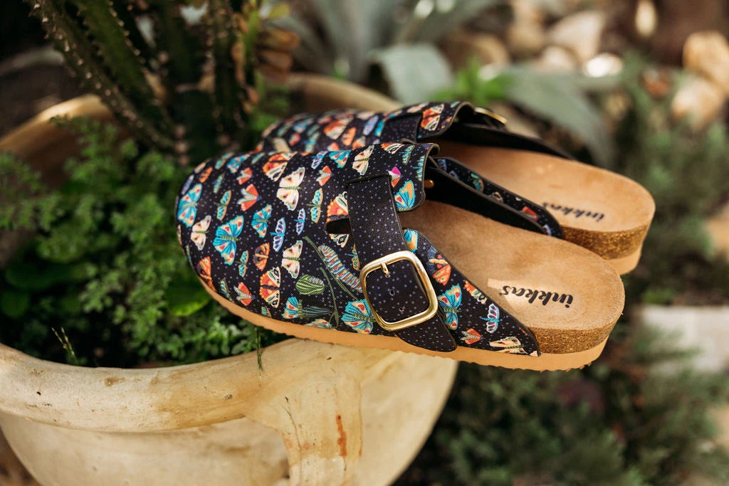 A pair of slip-on clog-style sandals with a multicolor fish pattern on a black background, resting on the edge of a rustic wooden planter. The sandals feature cork-look soles and a gold-tone buckle.