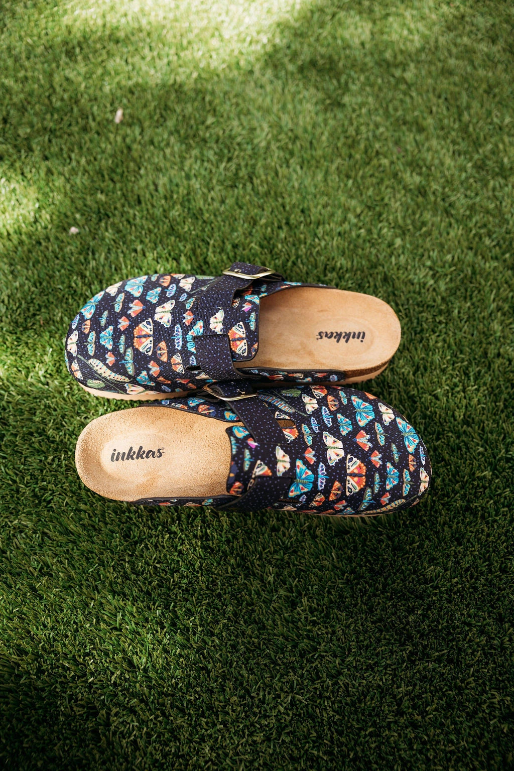 A pair of Inkkas slip-on clogs with a colorful geometric pattern on a dark navy base, placed on green grass.