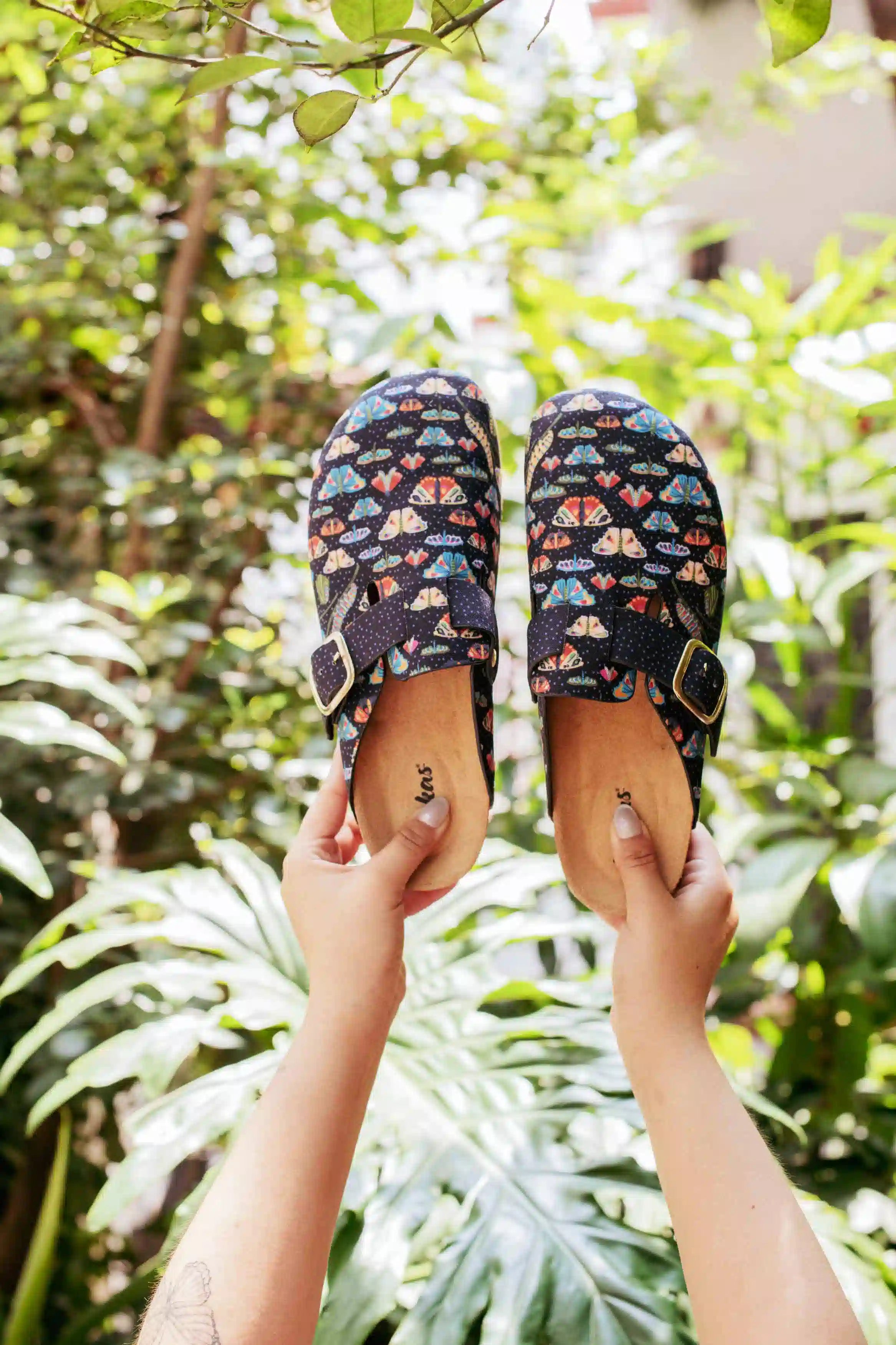 A pair of black clogs with a colorful tropical fish print and adjustable straps, held up by two hands against a lush green tropical foliage background.