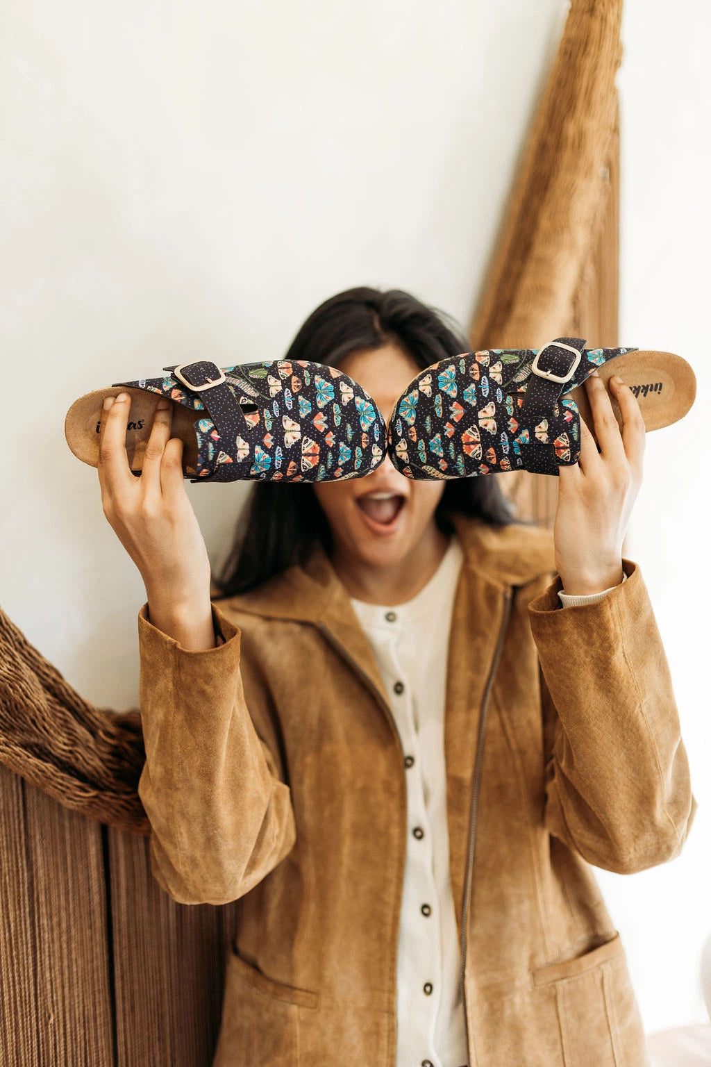 A woman wearing a tan suede jacket holds a pair of patterned slide sandals in front of her face, making a playful expression. The clogs feature a dark navy base with a colorful butterfly print in orange, blue, and white, complete with adjustable straps and light brown soles.