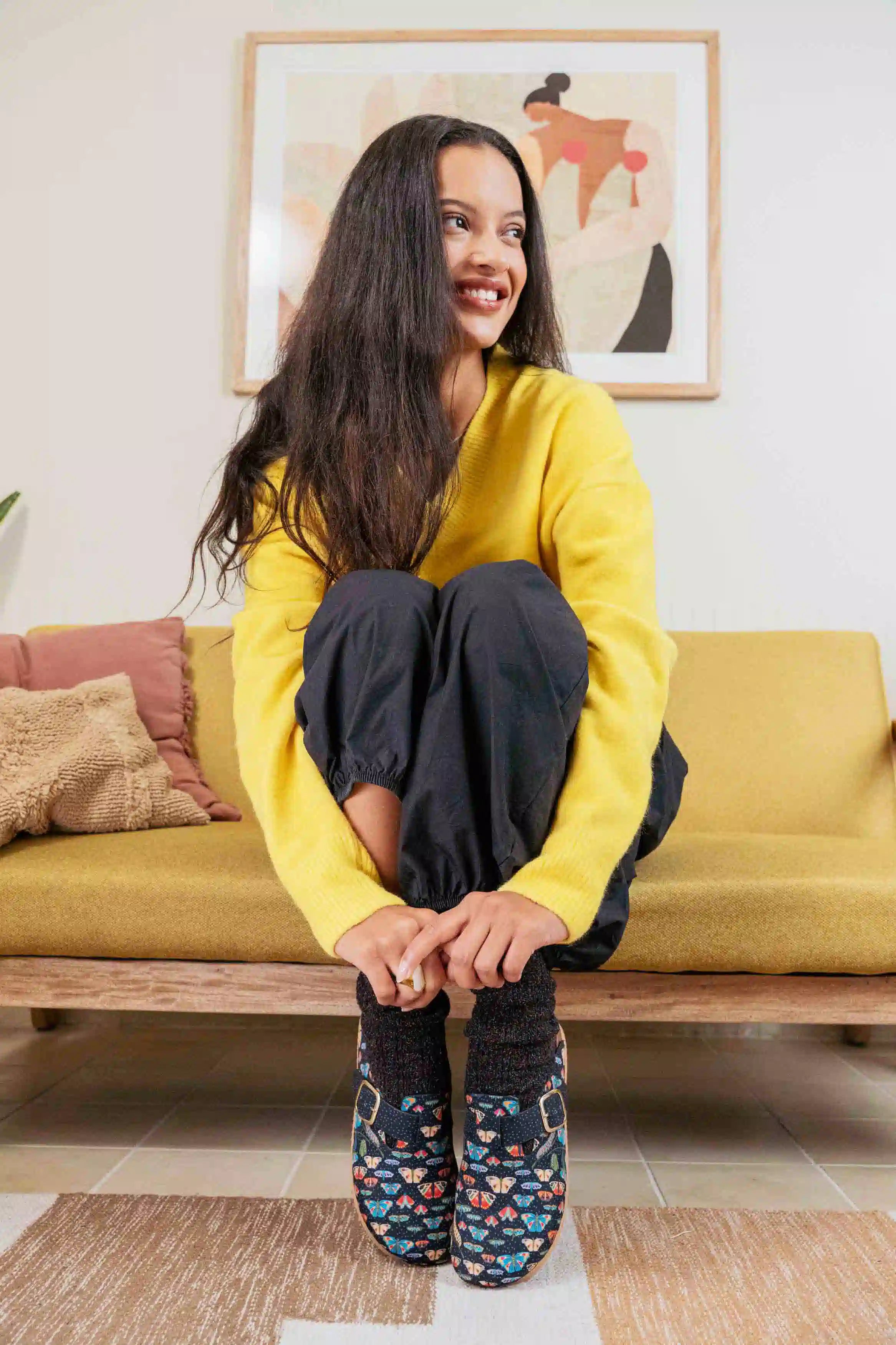 A woman sitting on a mustard yellow couch, wearing a bright yellow sweater, dark pants, Inkkas clogs and chunky socks. She is smiling and looking to the side.
