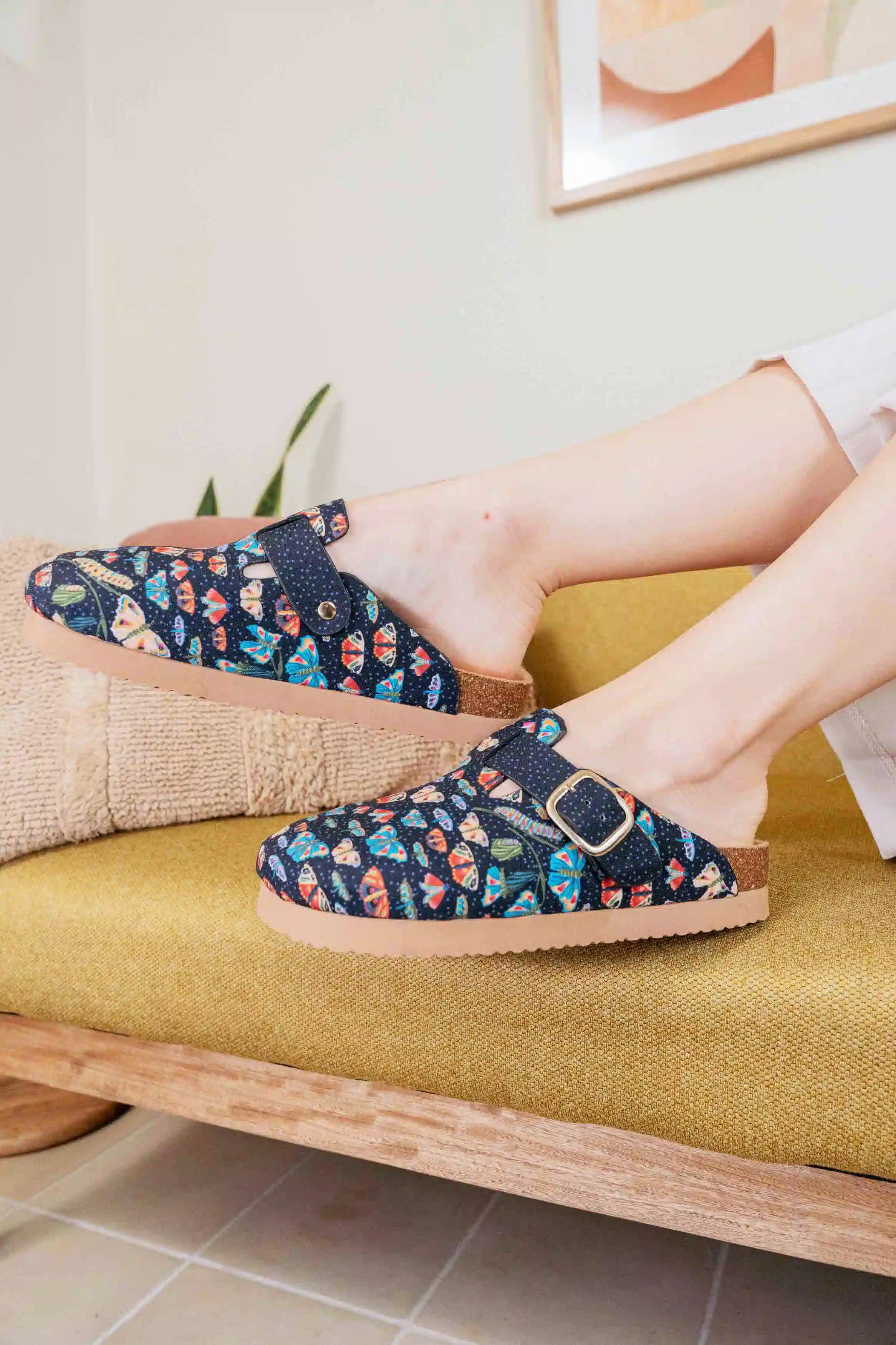 A person wearing a pair of slip-on clog-style slippers with a navy blue floral pattern and a gold-tone buckle, sitting on a mustard-yellow upholstered bench.