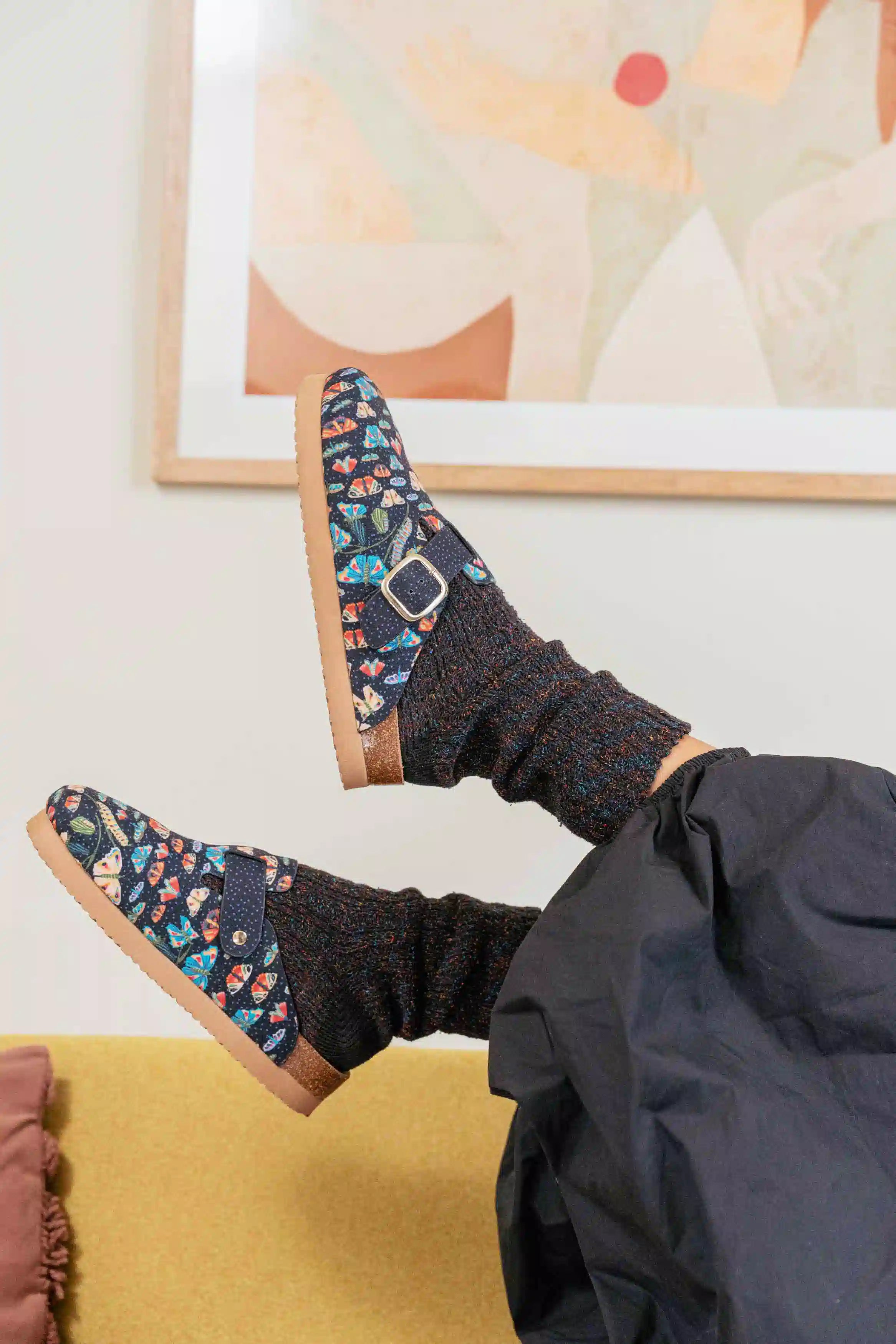 A person reclining indoors with legs raised, wearing dark patterned slip-on shoes with a buckle strap and dark knit socks, against a light wall with framed abstract art and a mustard-yellow surface below.