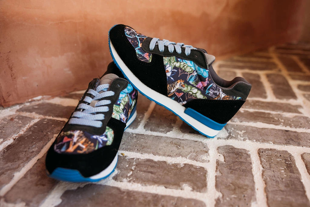A pair of black low-top sneakers with colorful graffiti-style printed panels, gray laces, and blue accents, resting on a brick floor against a terracotta wall.