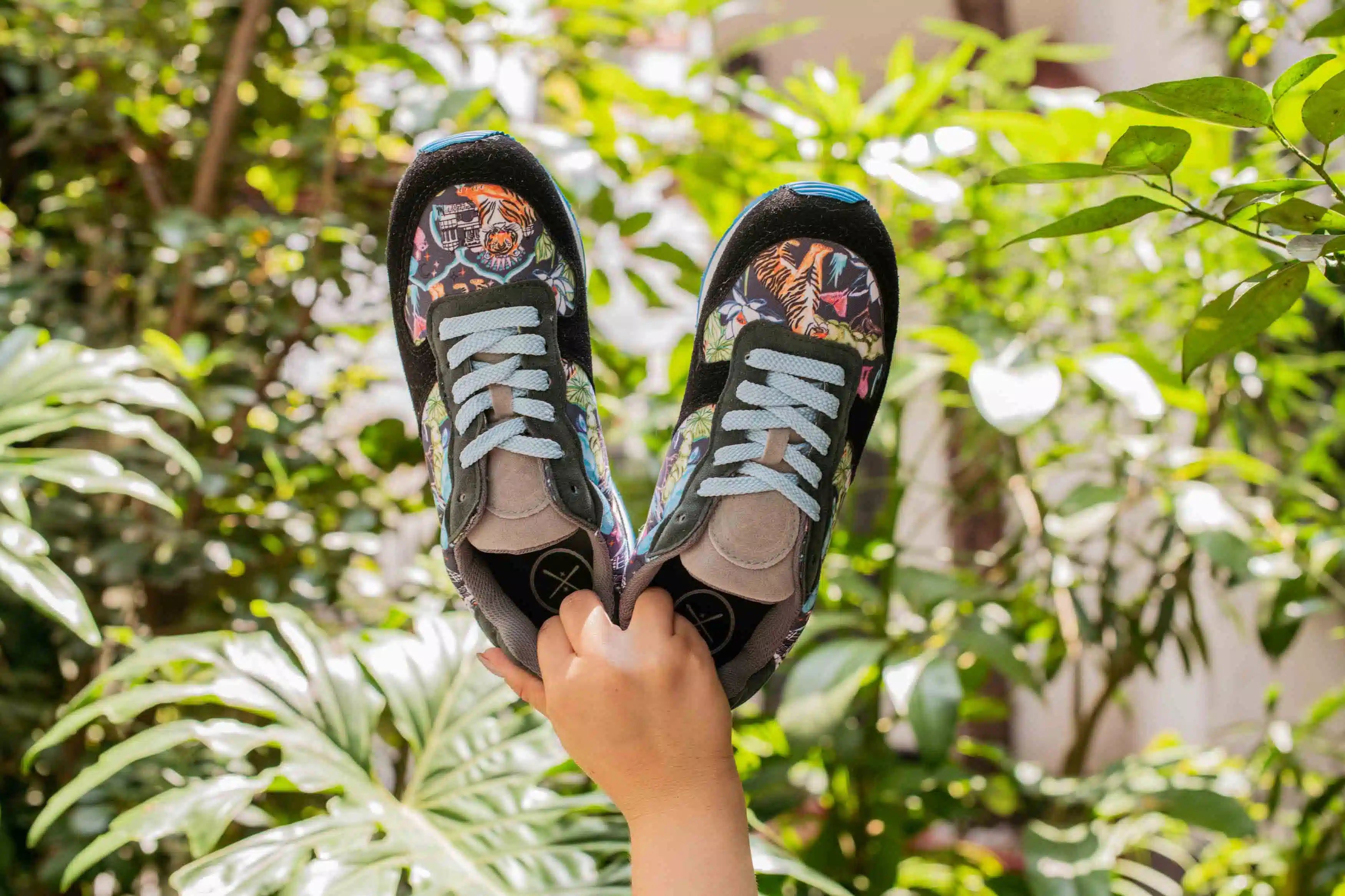 A pair of black low-top sneakers with colorful graffiti-style prints on the sides, held up by a hand against a lush green garden background.