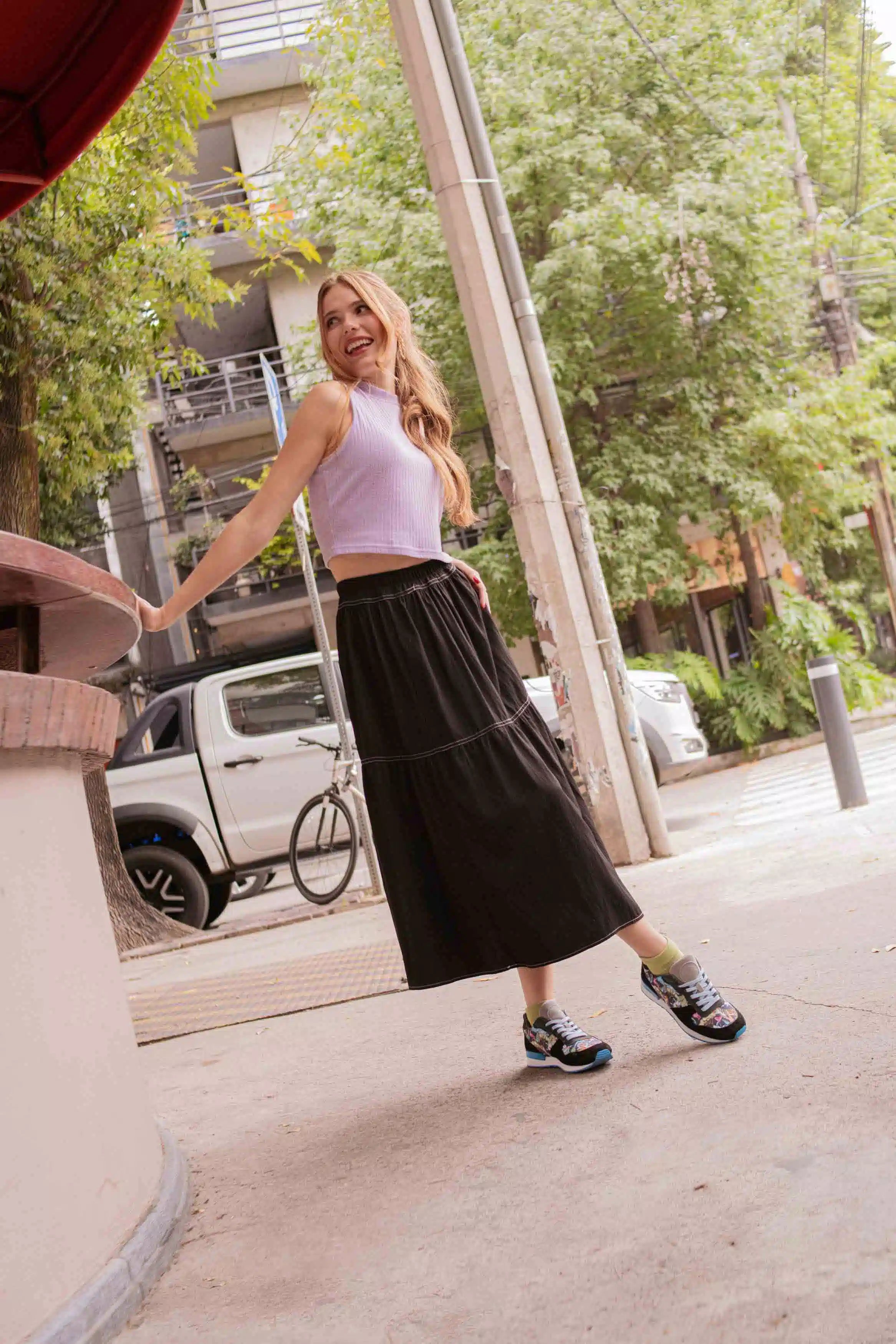 A woman posing outdoors on a city sidewalk, wearing a light lavender sleeveless crop top and a long black midi skirt with black sneakers.