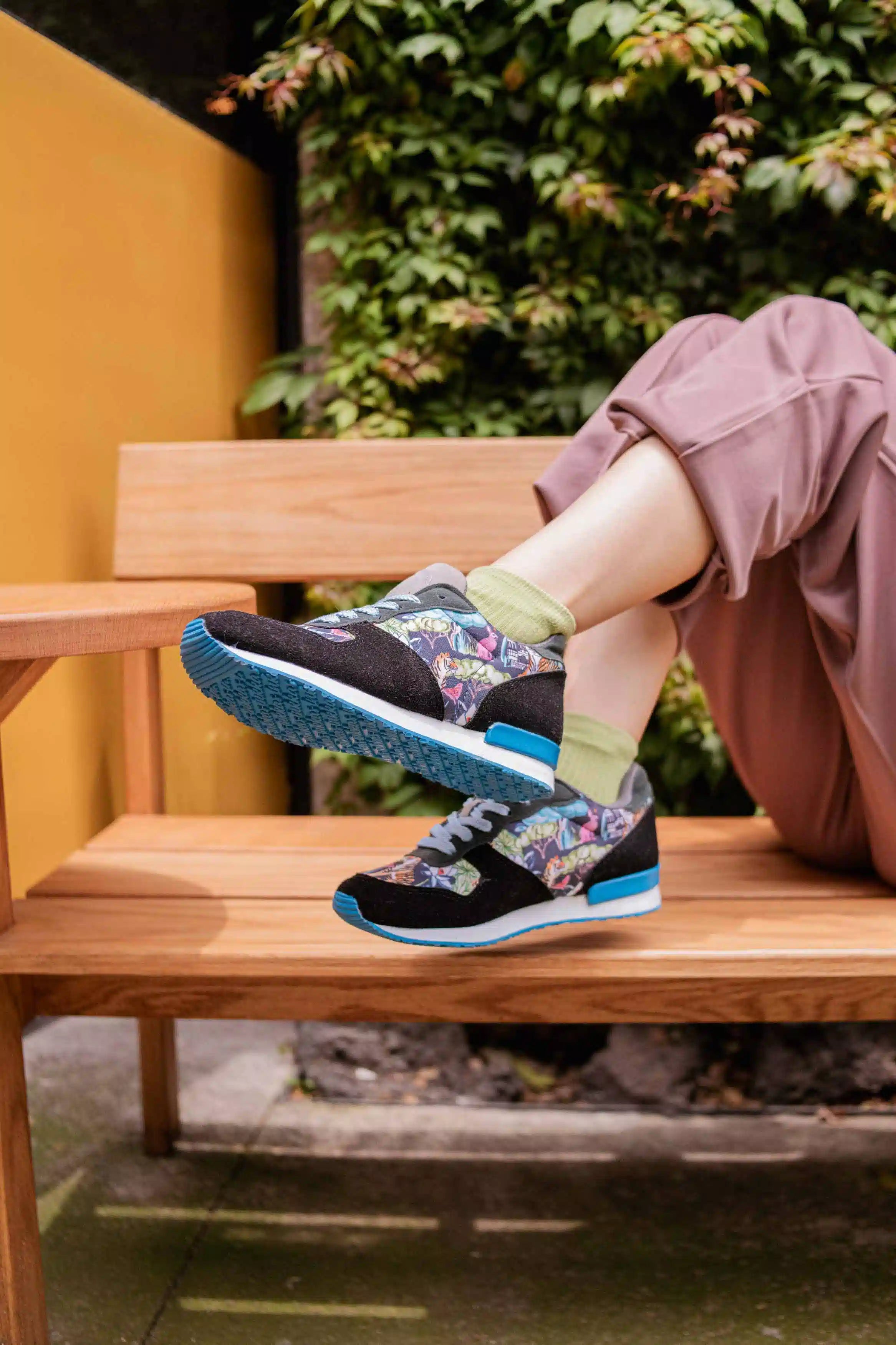 A person sitting on a wooden bench outdoors wearing black low-top sneakers with a colorful floral pattern on the sides, bright blue soles, and light green socks.