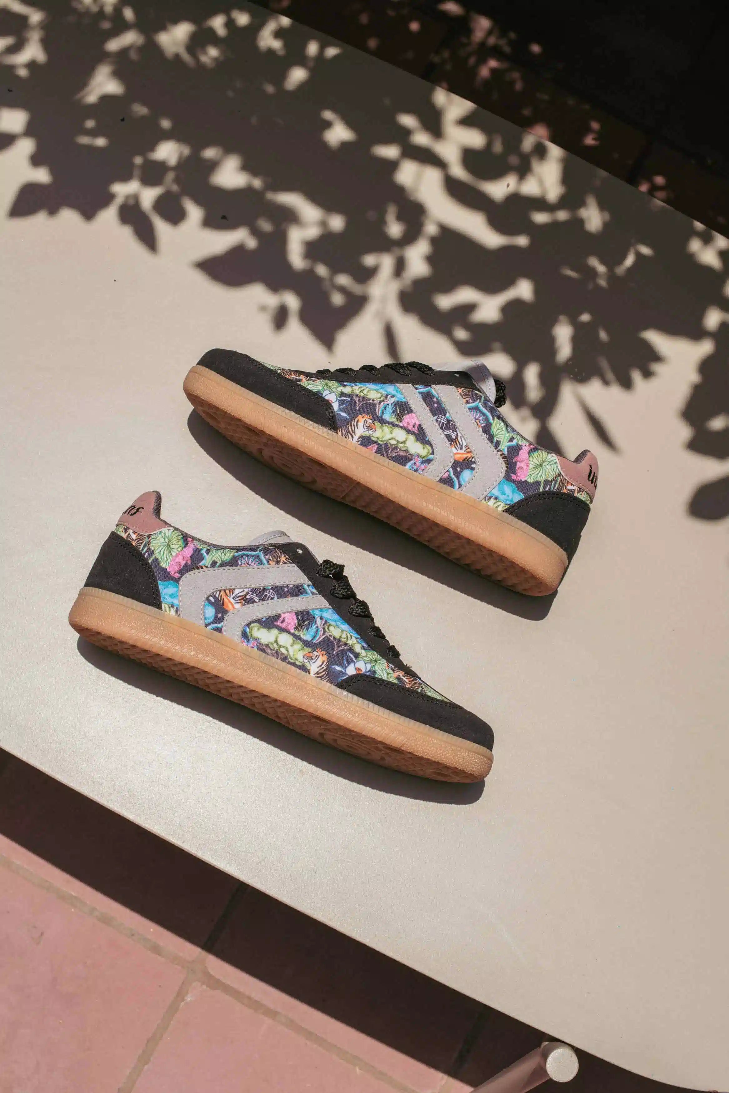 A pair of low-top sneakers with a colorful graphic pattern on the uppers, black toe and heel caps, and gum soles, resting on a light-colored outdoor table under leafy shadows.