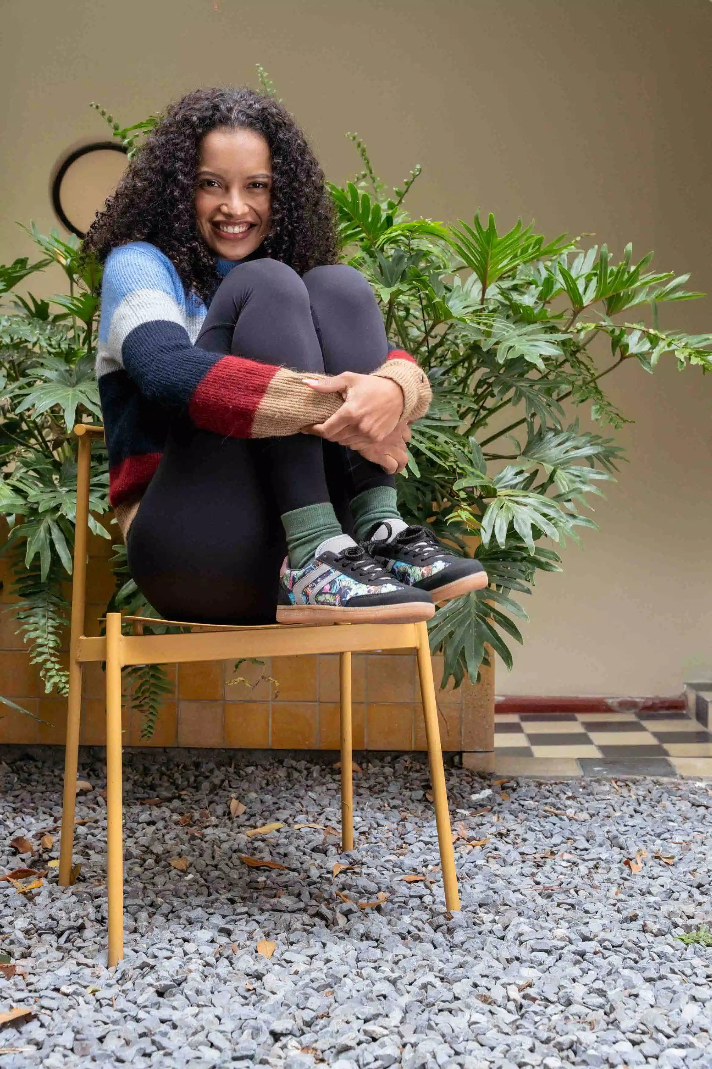 A smiling woman with curly hair sits cross-legged on a light wood chair in a garden courtyard. She is wearing a multicolor knit sweater, dark leggings, green socks, and patterned sneakers.