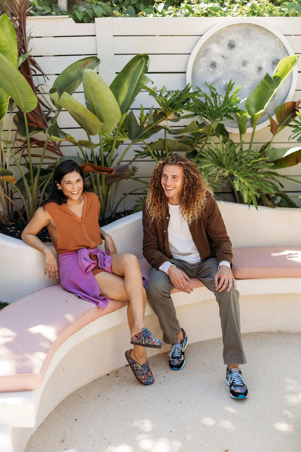 A man and a woman sit on a curved outdoor bench in a garden setting. The woman wears a rust-orange sleeveless top with Inkkas clogs, while the man wears a brown jacket over a white shirt, gray pants, and colorful sneakers.