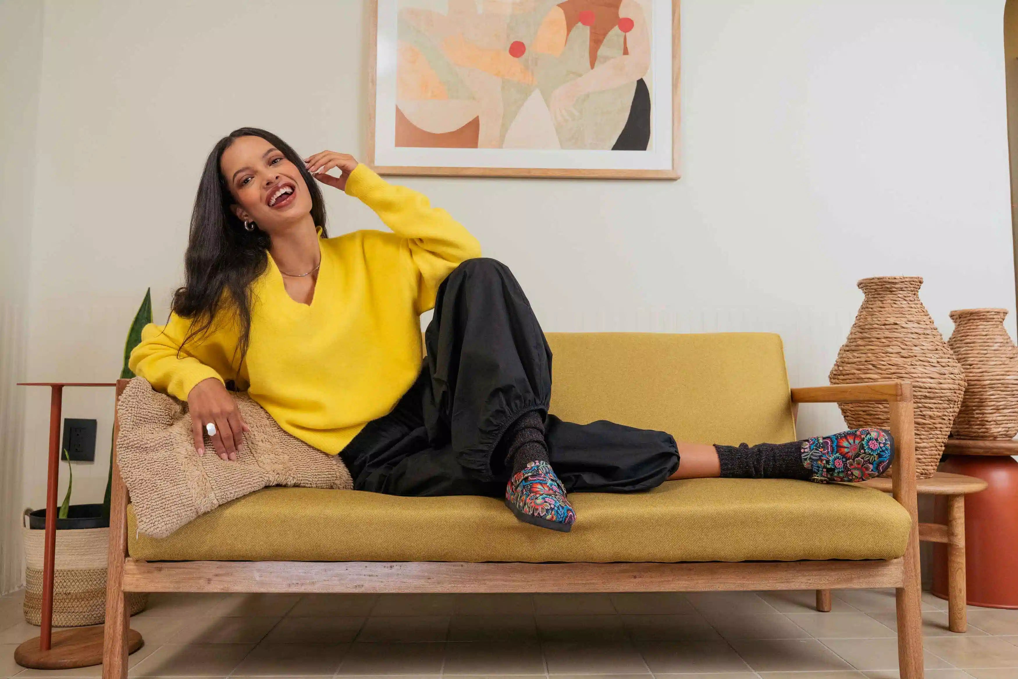 A woman reclining on a mustard yellow sofa in a modern living room setting, wearing a bright yellow sweater, black pants, Inkkas clogs and chunky socks.