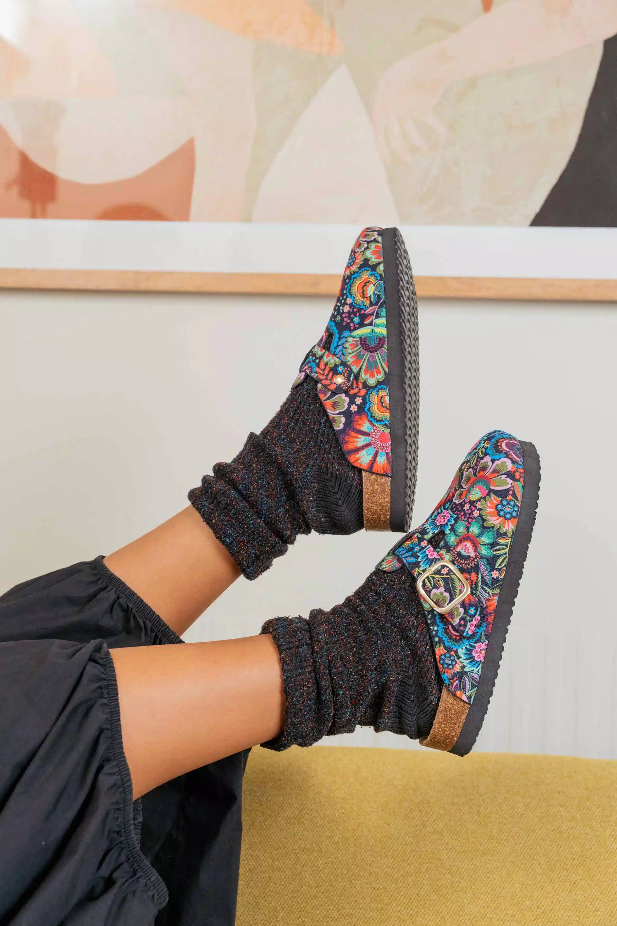A person wearing colorful floral slip-on mule shoes with black knit socks, shown from the knees down in a styled indoor setting.