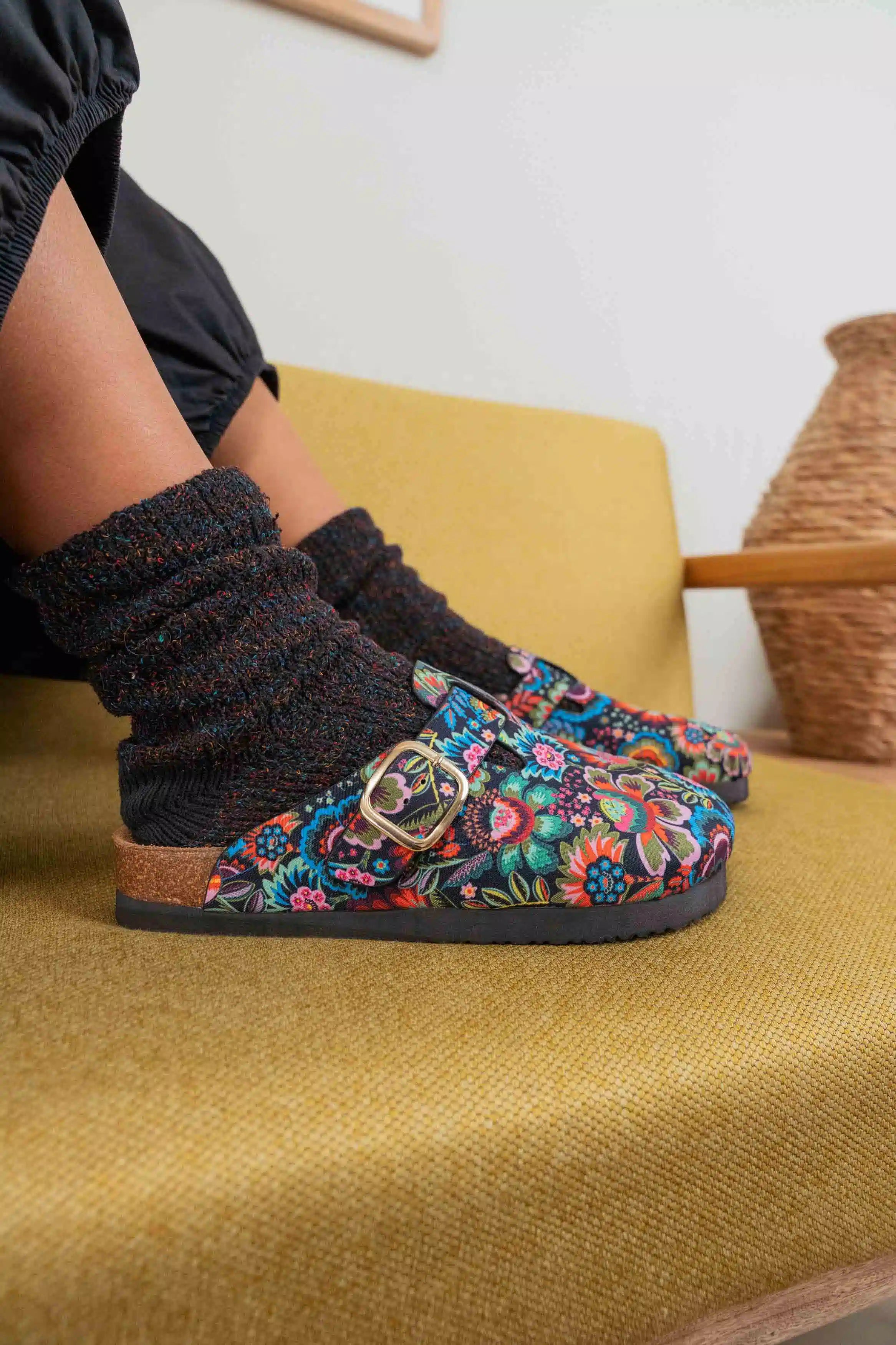 A person wearing colorful floral slip-on clogs with a gold buckle and black knit socks, sitting on a mustard yellow upholstered chair.