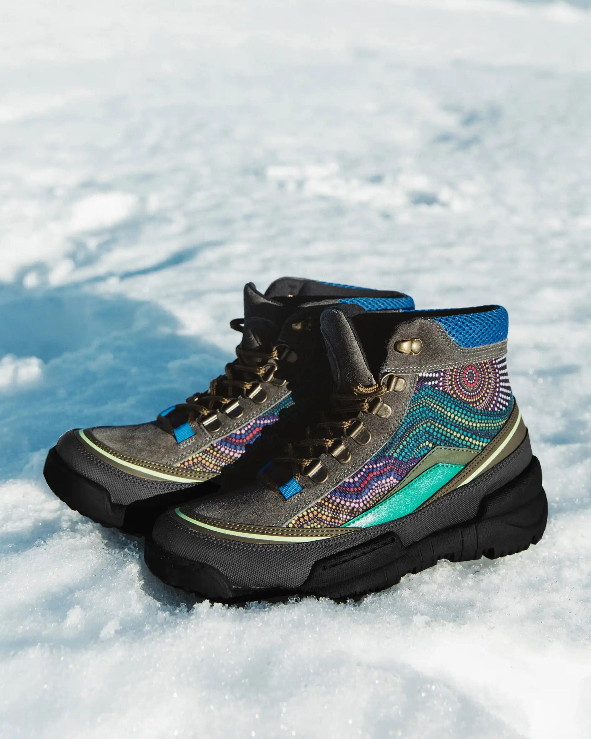 A pair of rugged lace-up hiking boots with black soles and gray uppers, decorated with colorful geometric beadwork and blue mesh panels, resting on a snowy white surface.