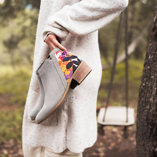 A hand holding the Inkkas Savannah Bootie, grey suede with a colourful floral panel, in woodland light