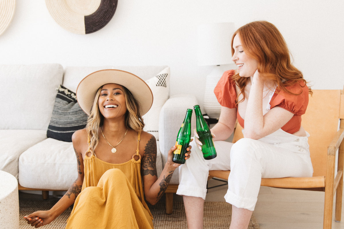 Two women sitting together in a sunlit room, laughing and clinking bottles, both wearing Inkkas