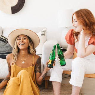Two women sitting together in a sunlit room, laughing and clinking bottles, both wearing Inkkas