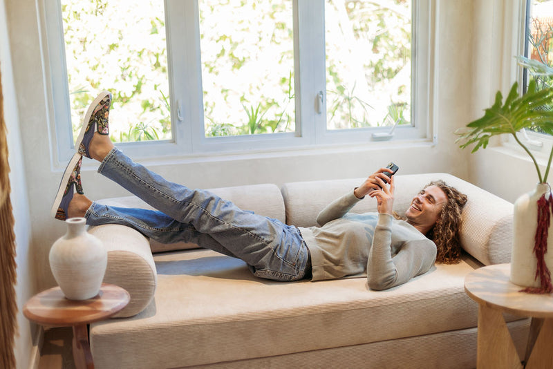 A woman lying on a sofa by a bright window, feet up, smiling at her phone