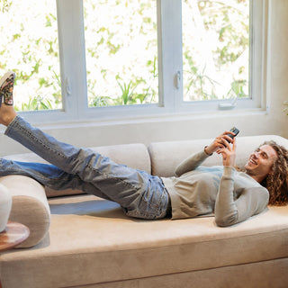 A woman lying on a sofa by a bright window, feet up, smiling at her phone