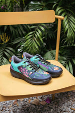 A pair of colorful athletic sneakers with a tropical floral and jungle print, displayed on a yellow wooden chair with a lush green leafy background.