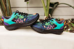 A pair of low-top athletic sneakers with a vibrant tropical floral print, resting on a white ledge with a blurred garden background.