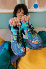 A person holds up a pair of colorful slip-on sneakers with a tropical underwater and sloth theme. The shoes feature bright turquoise, dark blue, green, and orange illustrations, with the word 'SLOTH' printed vertically on the side.