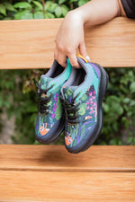 A person's hand holding a pair of colorful outdoor sneakers with a tropical jungle print featuring sloths and colorful flowers, set against a wooden bench and green foliage background.
