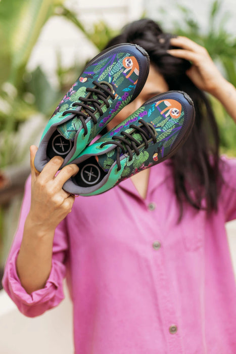 A person in a pink shirt holds up a pair of colorful sneakers with a tropical floral pattern featuring sloths and green leaves. The background is a soft-focus outdoor setting with green foliage.