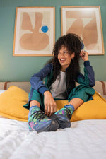 A woman sits on a bed wearing a matching teal and navy tracksuit, a gray t-shirt, and colorful patterned sneakers. She is posed in front of two framed abstract art prints on a sage green wall.