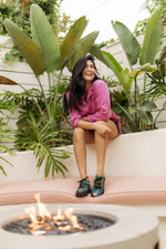 A woman sits on a curved outdoor bench in a garden setting, wearing a pink long-sleeve shirt and colorful patterned sneakers. A fire pit is visible in the foreground, with large green tropical plants behind her.