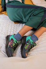A person reclining on a bed wearing green tracksuit pants and colorful athletic sneakers with blue socks. The sneakers feature a vibrant tropical leaf print in green, pink, and purple on a teal and black base.