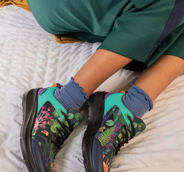 A person reclining on a bed wearing green tracksuit pants and colorful athletic sneakers with blue socks. The sneakers feature a vibrant tropical leaf print in green, pink, and purple on a teal and black base.