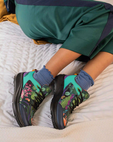 A person reclining on a bed wearing green tracksuit pants and colorful athletic sneakers with blue socks. The sneakers feature a vibrant tropical leaf print in green, pink, and purple on a teal and black base.