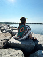 Person sitting on rocks by a body of water, wearing a colorful patterned blanket and hat.