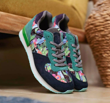 A pair of Inkkas joggers sneakers with a dark navy suede base, teal green overlays, and a colorful floral pattern on the side panels, resting on a wooden surface in a home setting.